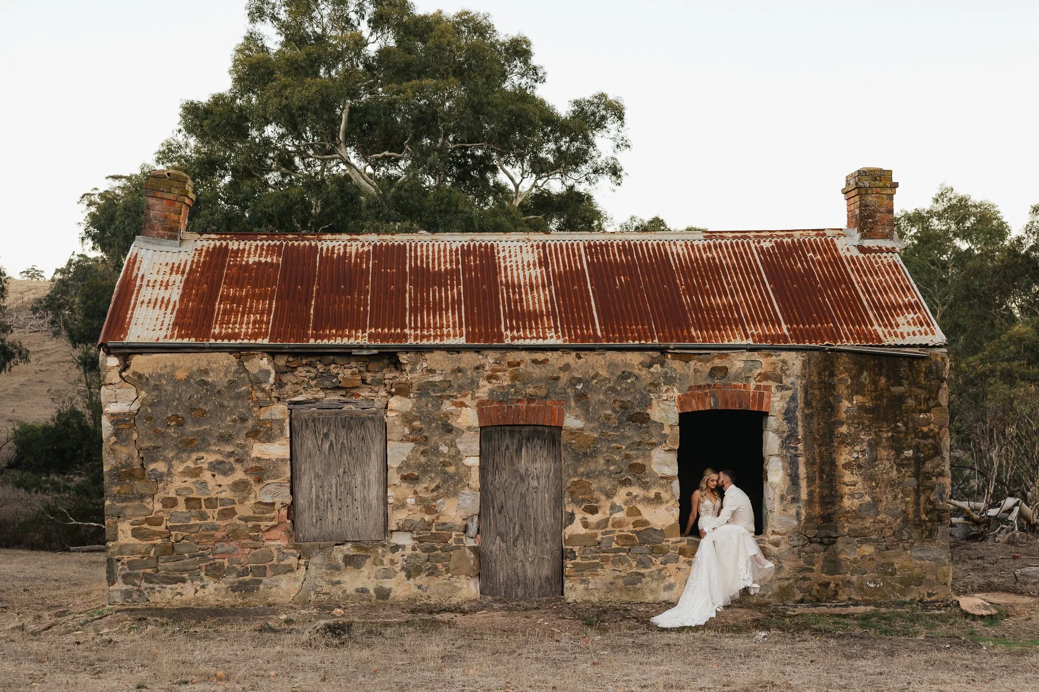 A bride and groom in wedding attire sitting in the doorway of an old, rustic stone building with a rusted metal roof, set in a barren landscape with trees in the background.