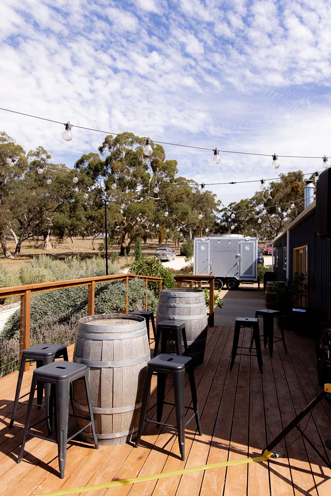 Outdoor patio with wooden deck, two large wine barrels used as tables, black bar stools, string lights overhead, trees and a blue sky in the background, and a food trailer in the distance.