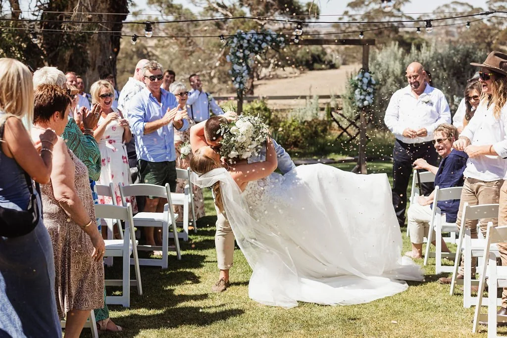 Bride and groom kissing at outdoor wedding ceremony, surrounded by guests clapping and celebrating, with string lights overhead.