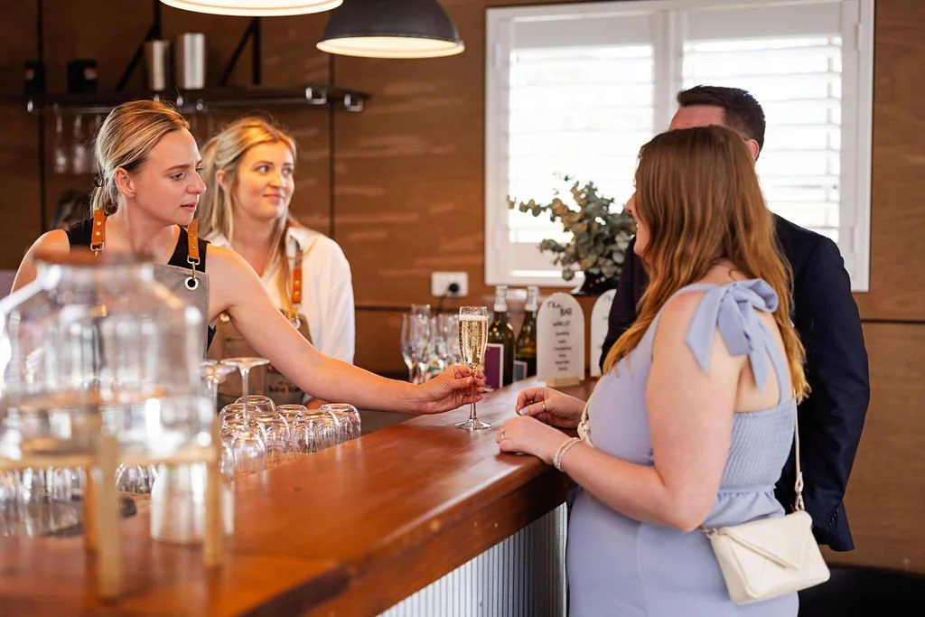 A bartender is handing a glass of champagne to a woman with red hair, while two other women stand nearby at a bar in a cozy, well-lit restaurant or café.
