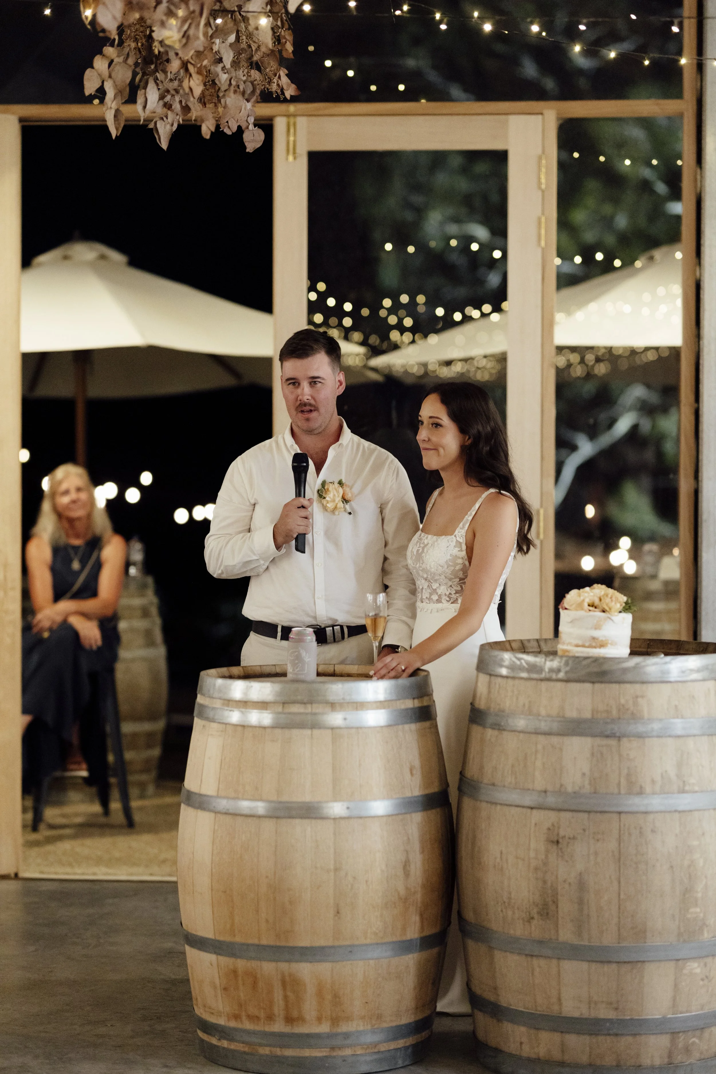 A couple giving a wedding speech at their reception, with a man holding a microphone and a woman standing next to him, standing behind two wine barrels, with a wedding cake on one of the barrels. The background shows string lights and outdoor umbrell
