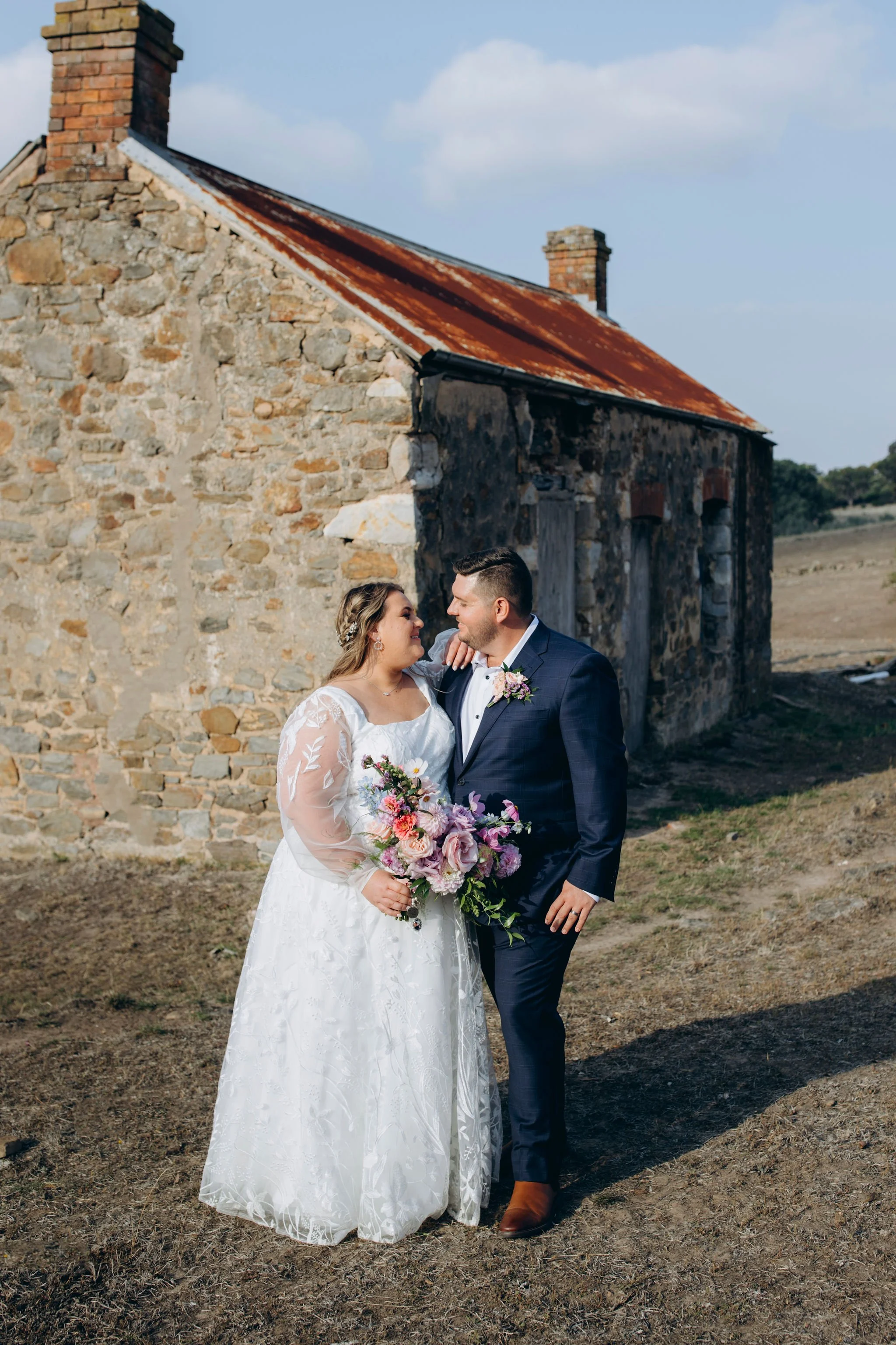 A bride and groom in wedding attire standing outdoors in front of a rustic stone building with a rusty metal roof, looking at each other affectionately, holding a bouquet of pink, purple, and white flowers.