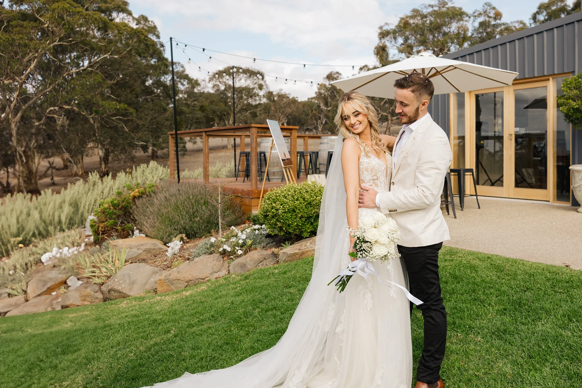 A bride and groom smiling and holding each other outdoors, with the bride holding a bouquet of white roses, in a garden area near a building with an umbrella and string lights overhead.