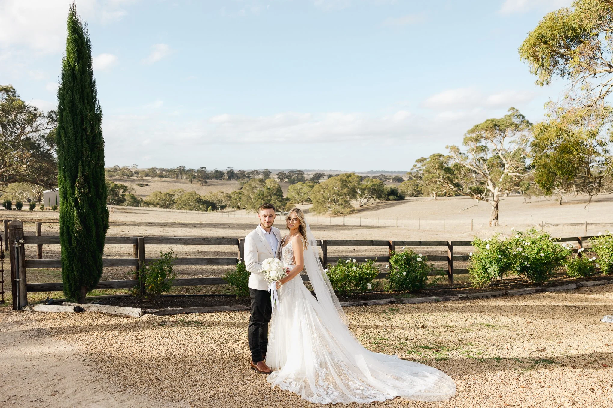 A bride and groom in wedding attire standing outdoors in a rural landscape with open fields, trees, and a fence, under a partly cloudy sky.
