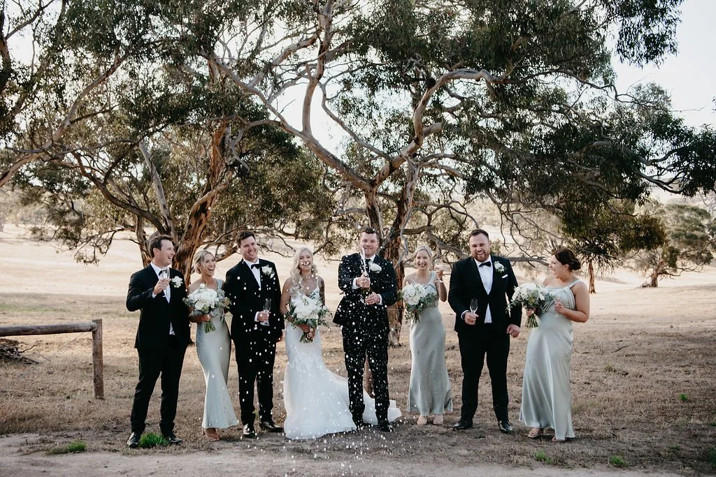 A wedding party with seven people, including bride and groom, celebrating outdoors under a large tree, with confetti in the air.