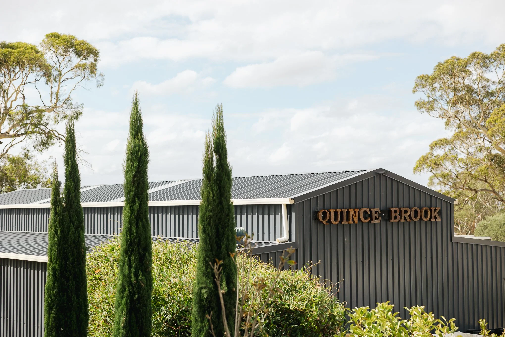 A modern gray metal building with the sign 'Quince Brook' on the front, surrounded by tall cypress trees and other greenery, under a partly cloudy sky.