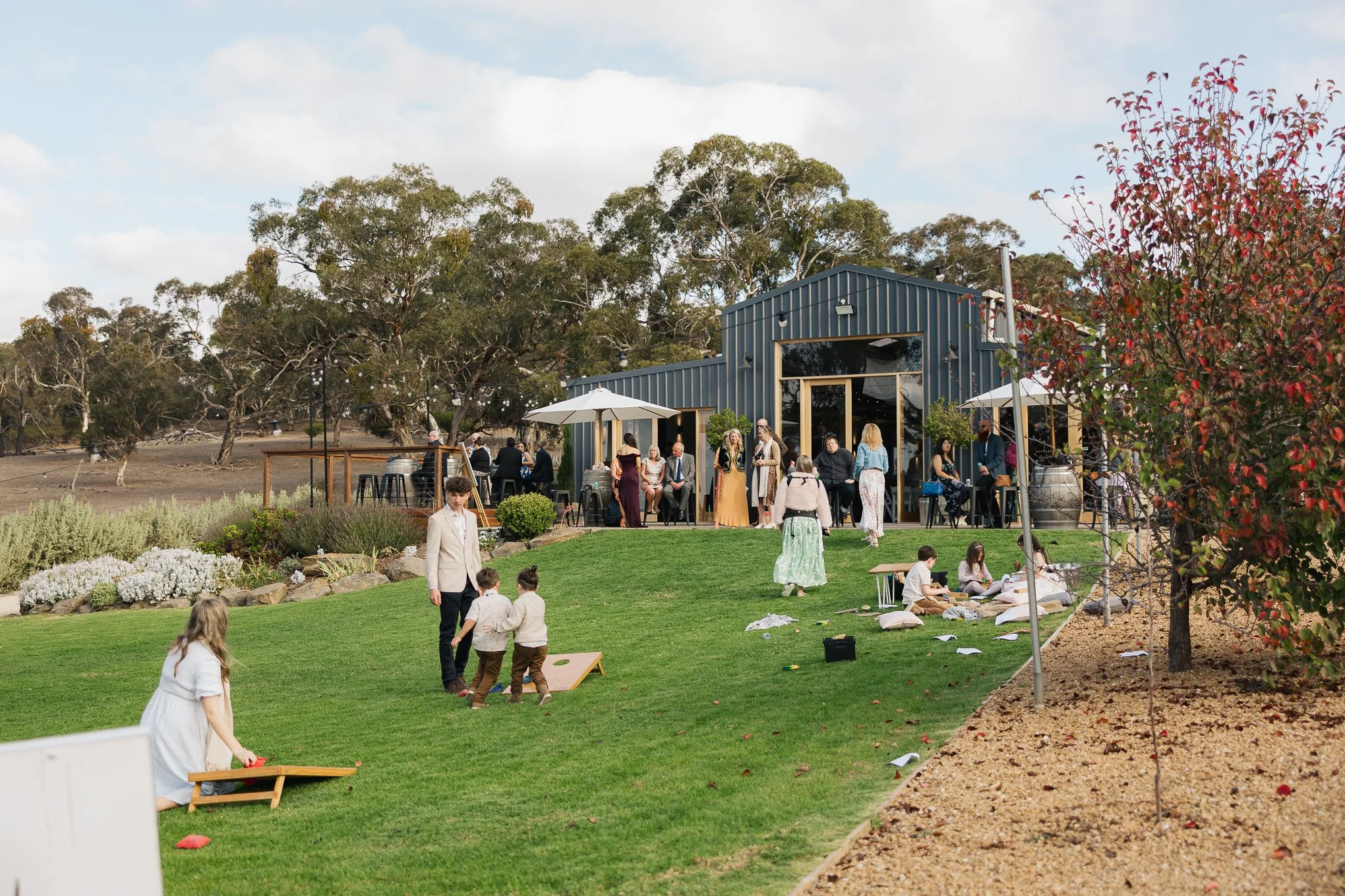 Outdoor gathering at a rustic venue with people socializing, children playing on grass, and some seated under umbrellas near a blue barn surrounded by trees and landscaped areas.
