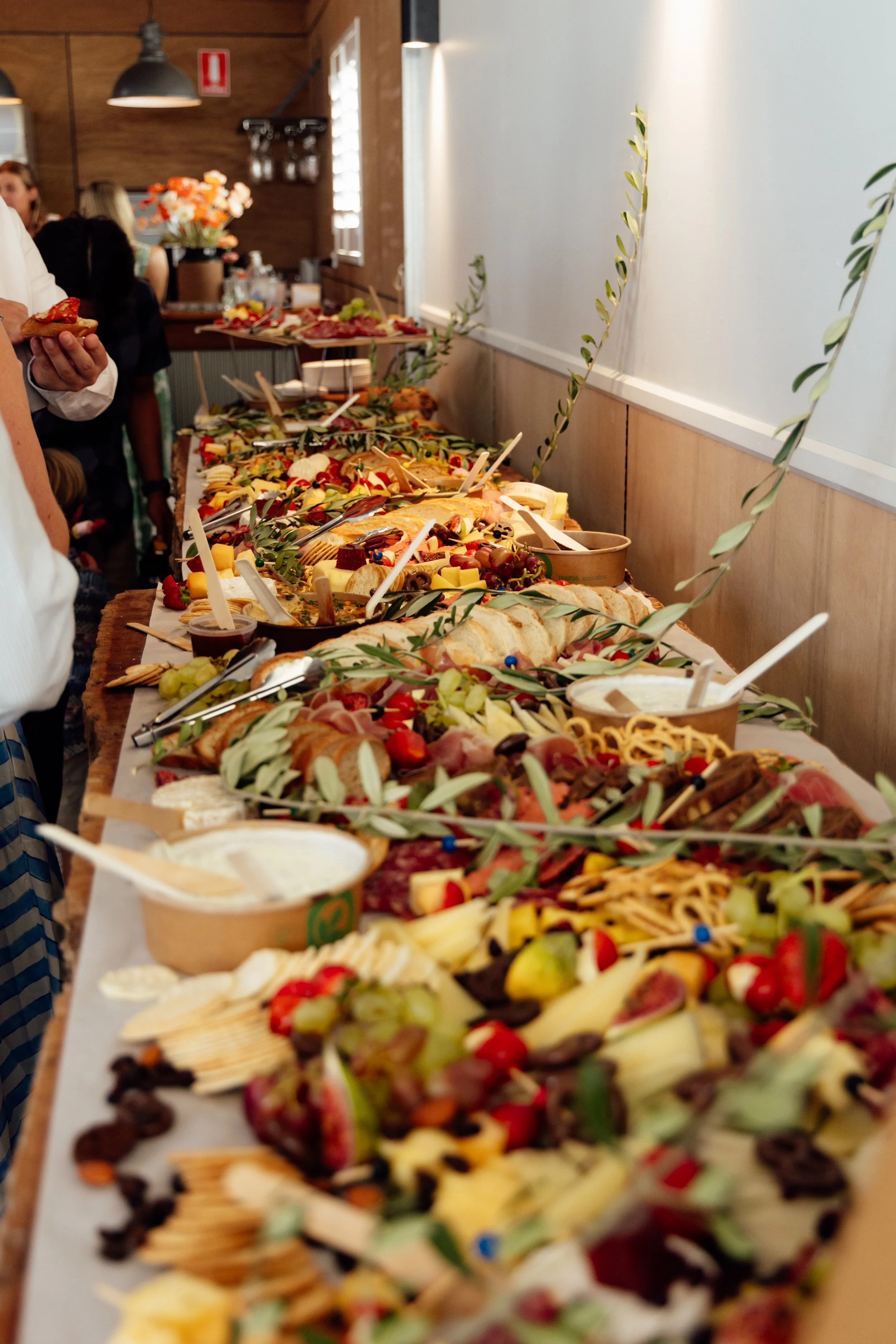A long table filled with an assortment of food including cheese, meats, grapes, bread, and crackers, decorated with olive branches in a buffet style setting at a social gathering.