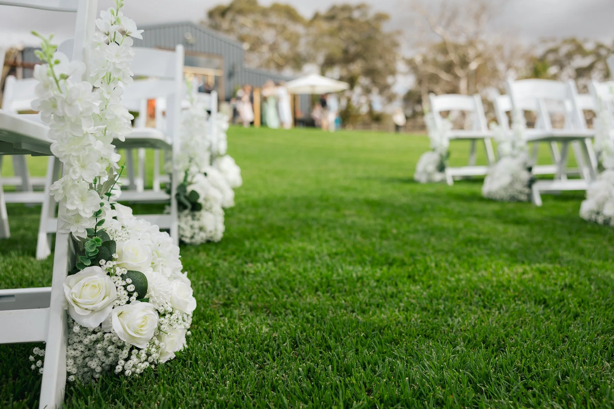 Outdoor wedding ceremony setup with white chairs decorated with white flowers on green grass, with a building and trees in the background.