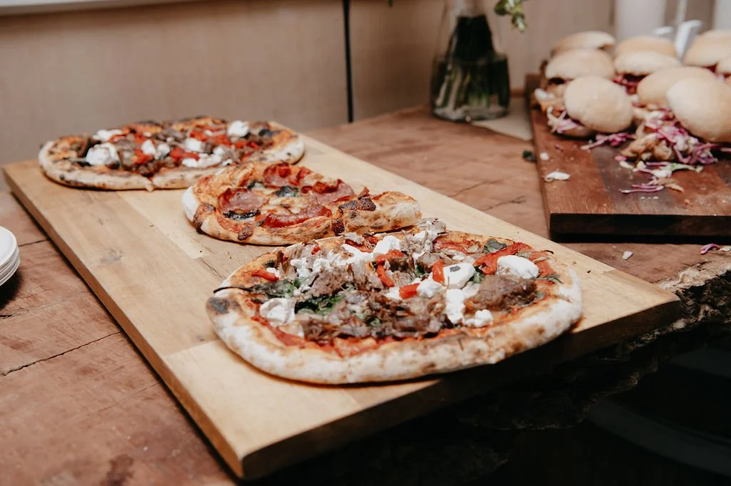 Three pizzas on a wooden serving board, with two other pizzas and bread rolls in the background.