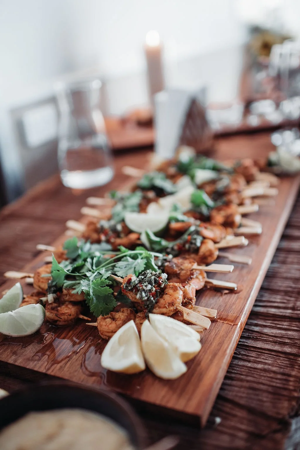 A wooden serving board with skewered grilled shrimp garnished with cilantro, lime wedges, and a dollop of sauce, on a dining table with glasses of water in the background.