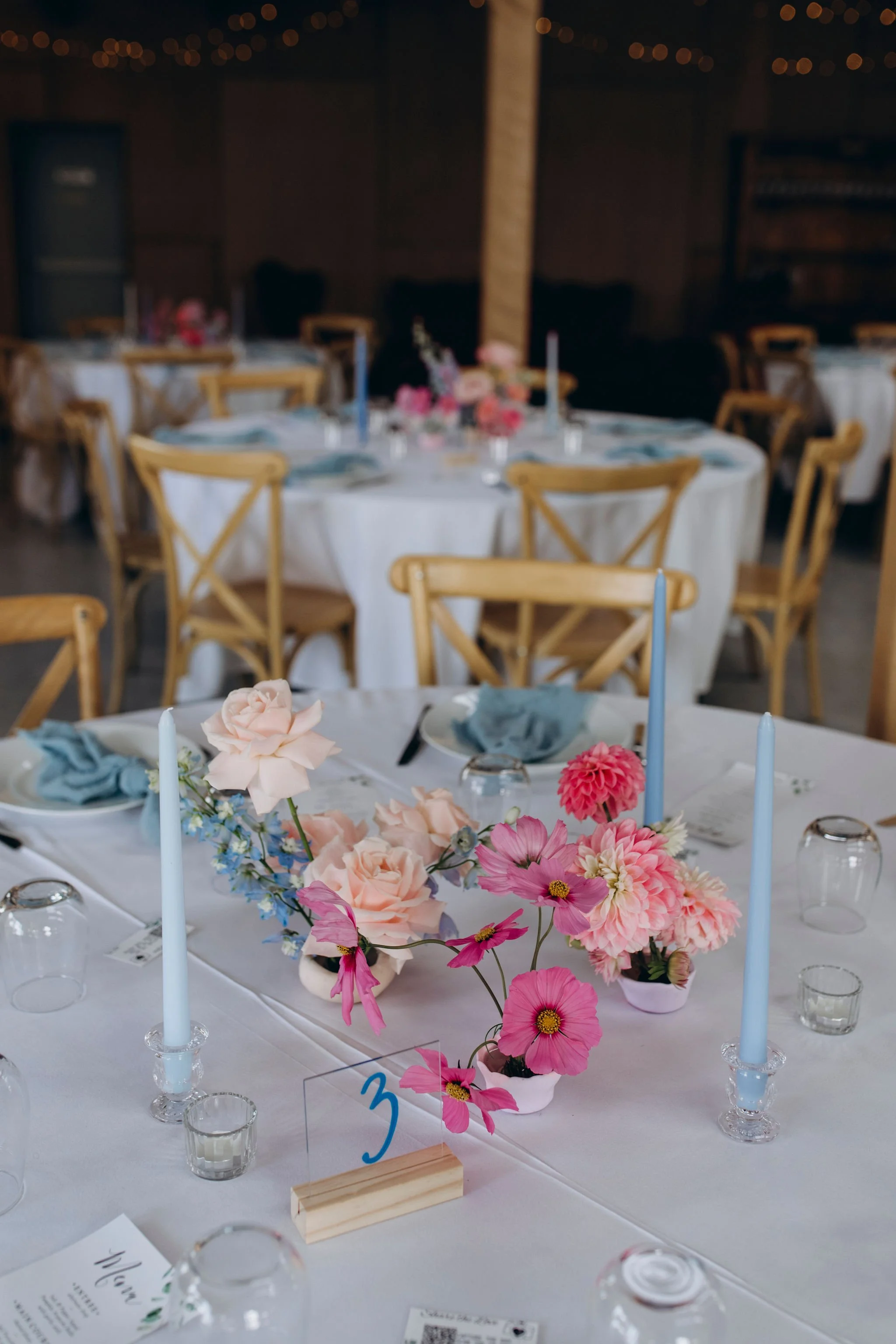 A decorated event table with pink, white, and blue flowers, tall blue candles, glasses, and place settings in a rustic indoor venue.