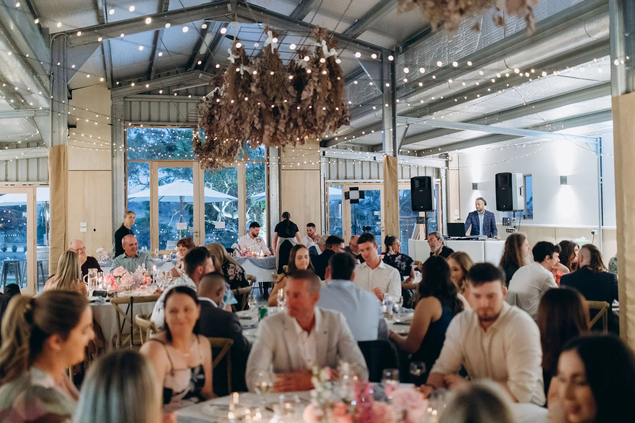 A bustling indoor event with guests seated at round tables, soft lighting, and a DJ on a platform. Outside, large windows reveal a patio with umbrellas and trees.