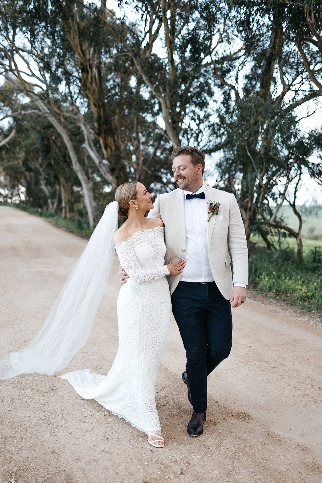 A bride and groom walk arm in arm along a dirt path surrounded by trees, smiling at each other.