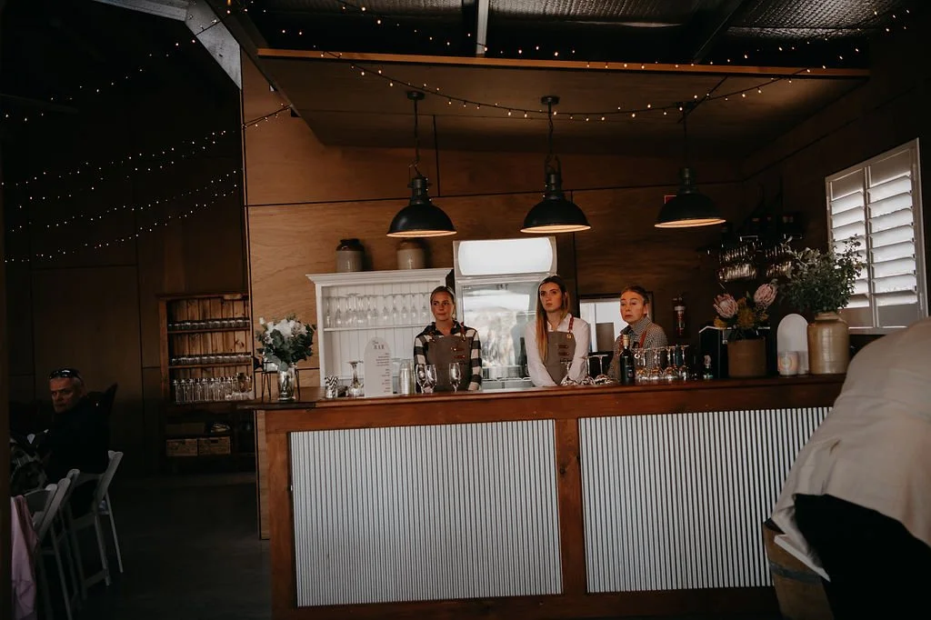 Three women behind a wooden bar counter with hanging pendant lights in a cozy, warmly lit interior space, decorated with flowers and bottles.