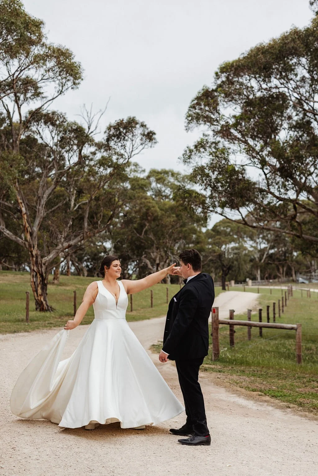 A bride in a white wedding gown and a groom in a black suit standing on a dirt path in a park with trees, with the bride touching the groom's nose playfully.
