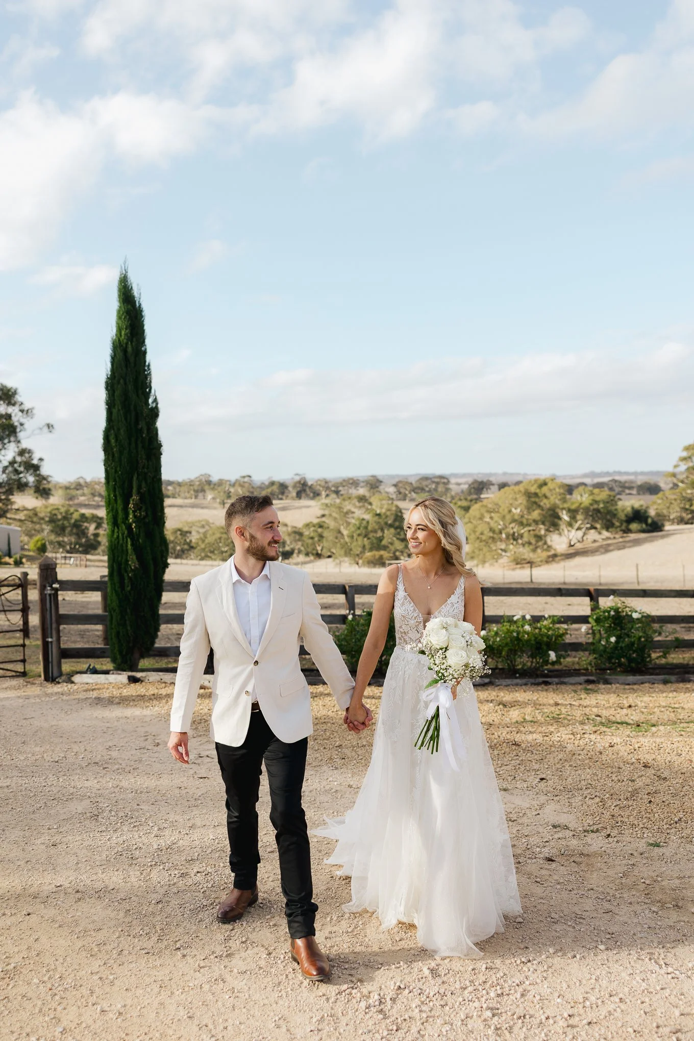 A bride and groom walking hand in hand outdoors in a rural setting with hills and trees, smiling at each other on their wedding day.