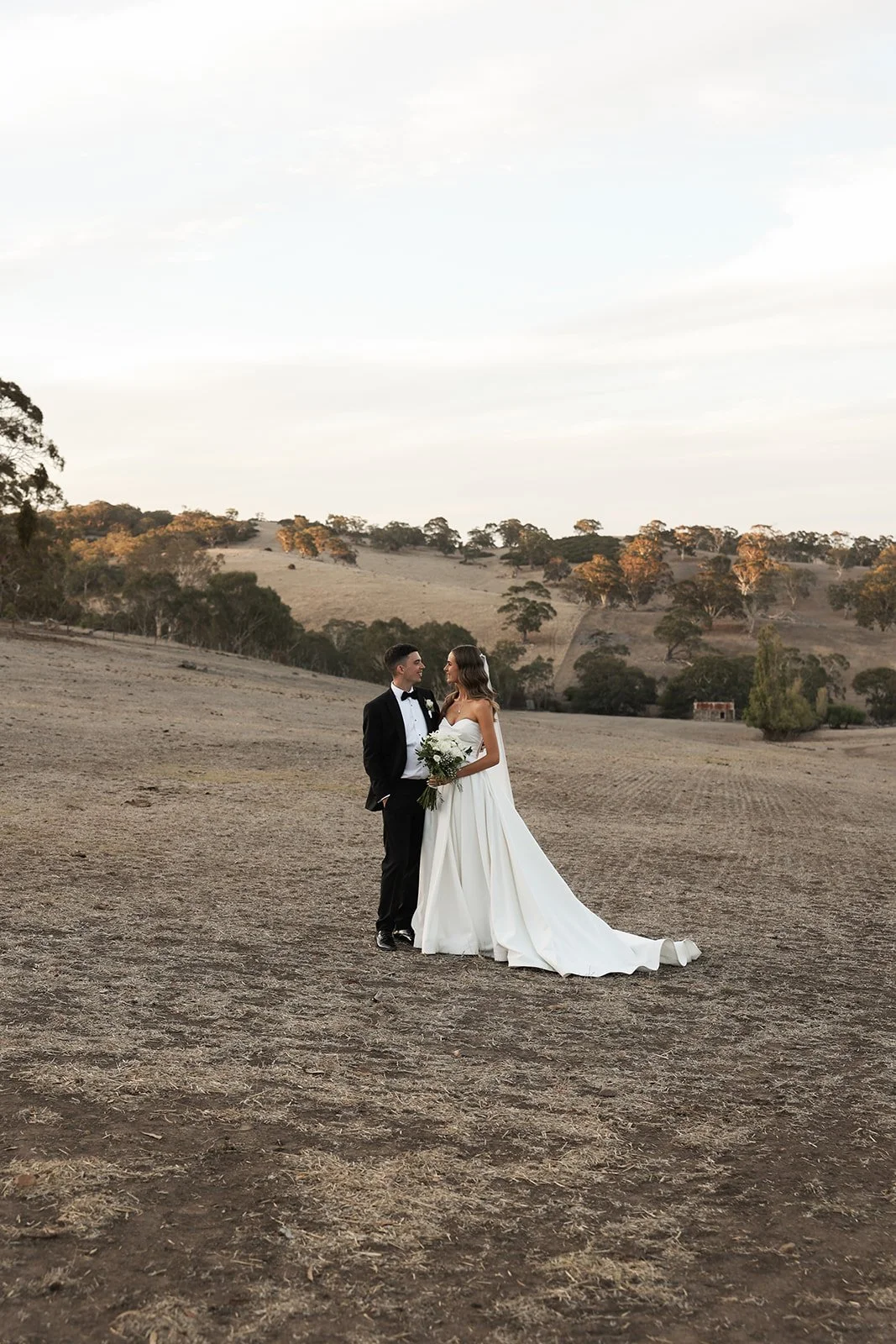 Bride and groom standing in a wide open, grassy field with rolling hills and scattered trees in the background, during sunset.