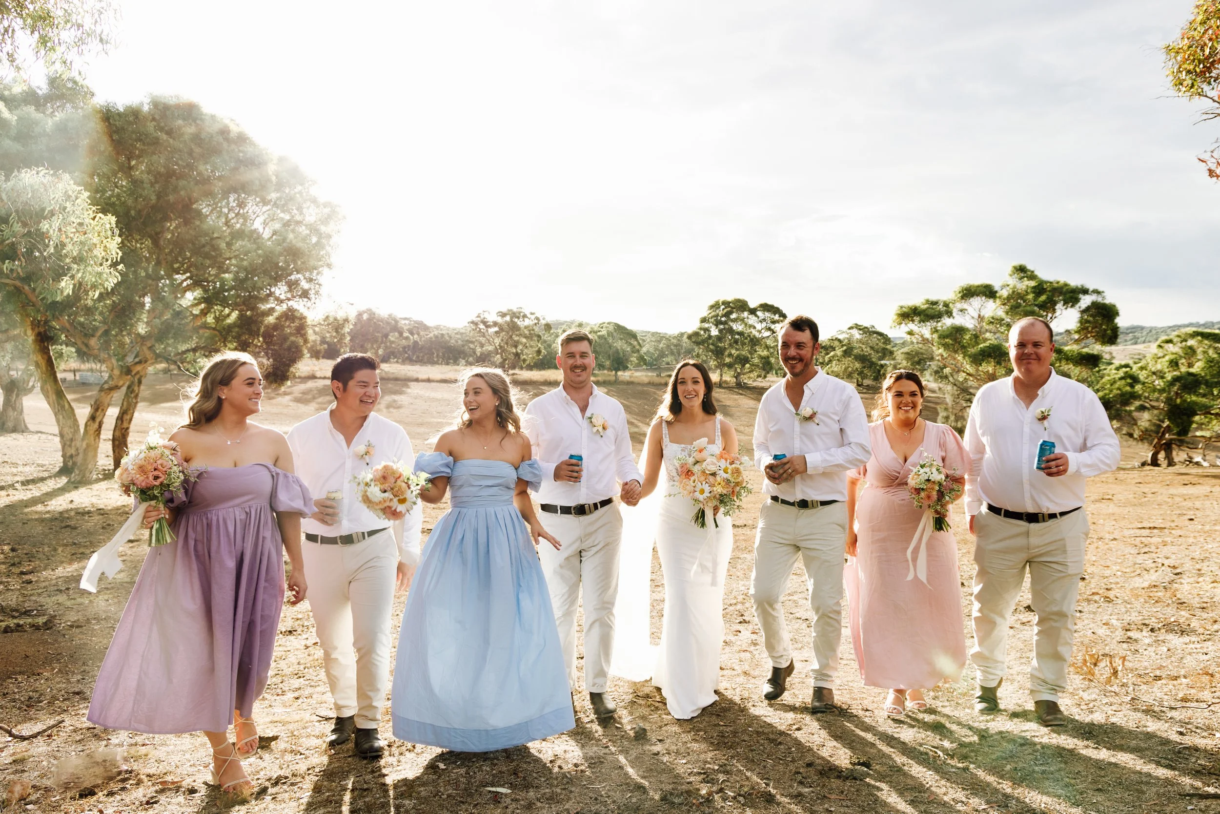 Group of eight people dressed in formal attire walking outdoors with trees and open landscape in the background, holding flowers and drinks, smiling and enjoying each other's company.