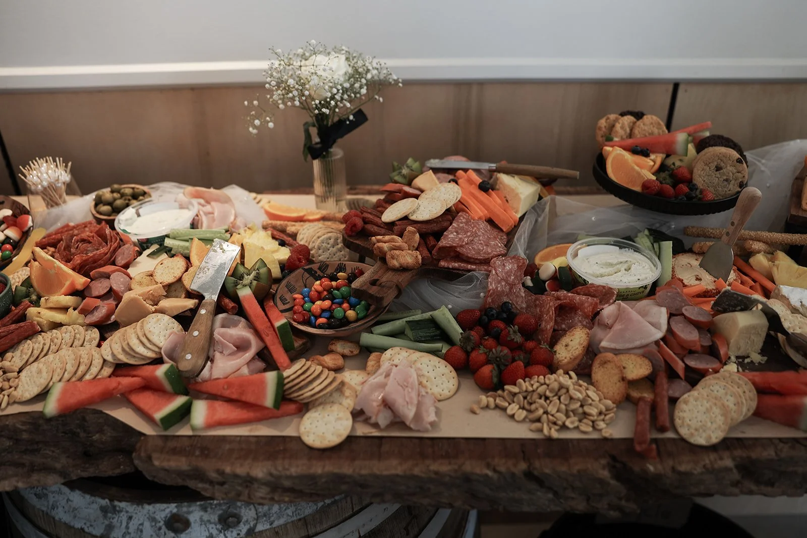 Charcuterie board with assorted meats, cheeses, crackers, fruits, vegetables, and candies, decorated with a small vase of white flowers.