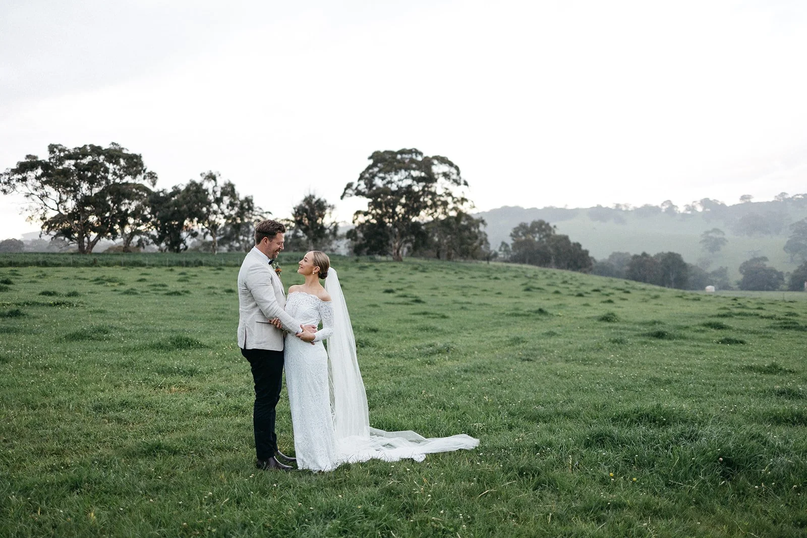 A bride and groom standing on a grassy field, gazing into each other's eyes, with trees and hills in the background during sunset.