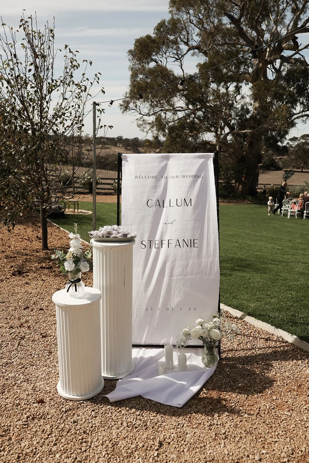 Wedding welcome sign with floral arrangements at an outdoor venue, featuring a white signboard with black text reading "Welcome to our wedding Callum and Steffanie" and the date "22 03 25."