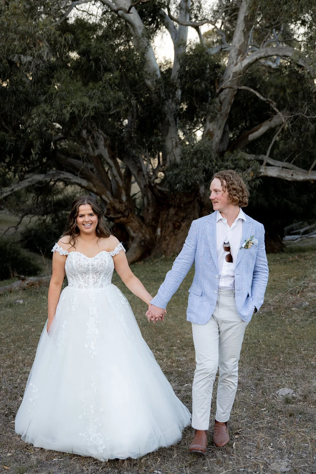 A newlywed couple walking hand in hand outdoors; the bride in a white ball gown with lace details, the groom in a light blue blazer, white shirt, and beige pants with sunglasses hanging from his shirt, against a large tree with a sprawling canopy.
