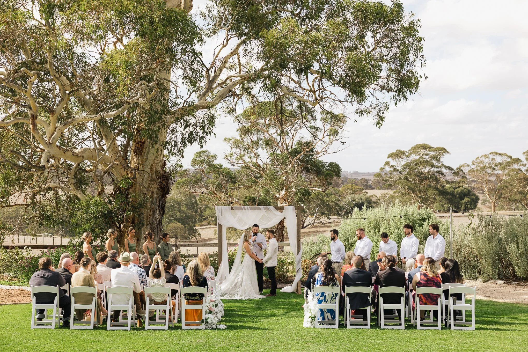 Outdoor wedding ceremony with bride and groom exchanging vows under a white draped arch, surrounded by seated guests, in a lush green garden with large trees and clear sky in the background.