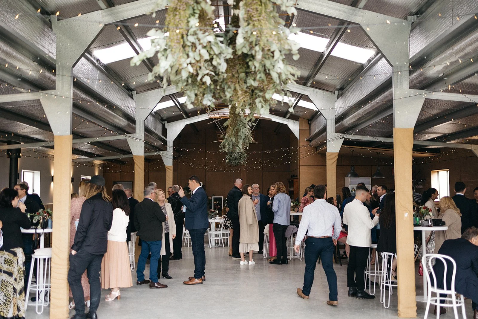 Guests mingling and chatting at a wedding reception in a decorated rustic venue with hanging floral arrangements and string lights.