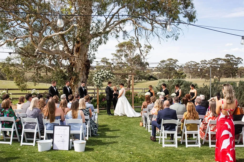 Outdoor wedding ceremony with a bride and groom exchanging vows under a wooden arch decorated with flowers, surrounded by guests seated on white chairs on a grassy area, with trees and a clear sky in the background.