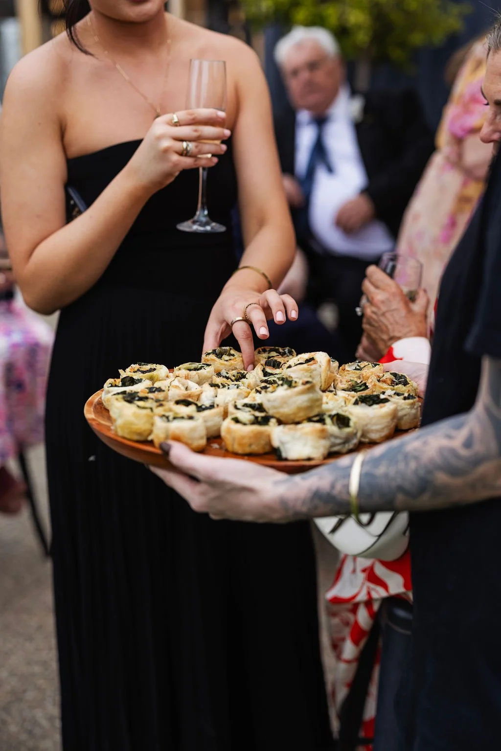 A woman in a strapless black dress holding a glass of champagne. She is reaching for some hors d'oeuvres from a tray with finger foods topped with black olives. Other guests are visible in the background at an outdoor event.