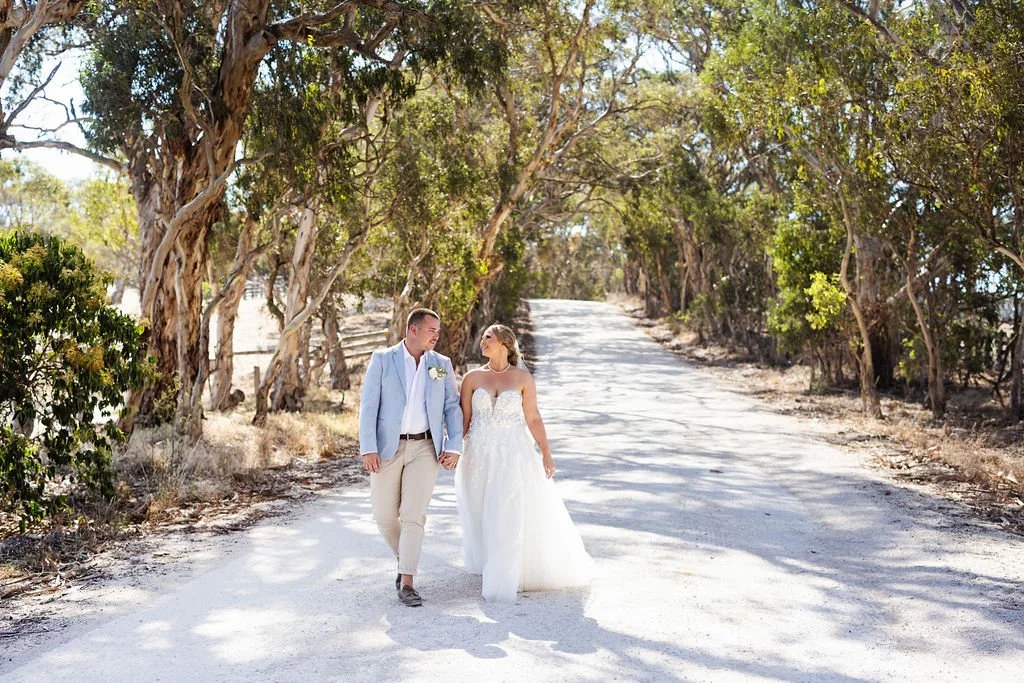 A newlywed couple walking hand in hand on a dirt road lined with trees on a sunny day.
