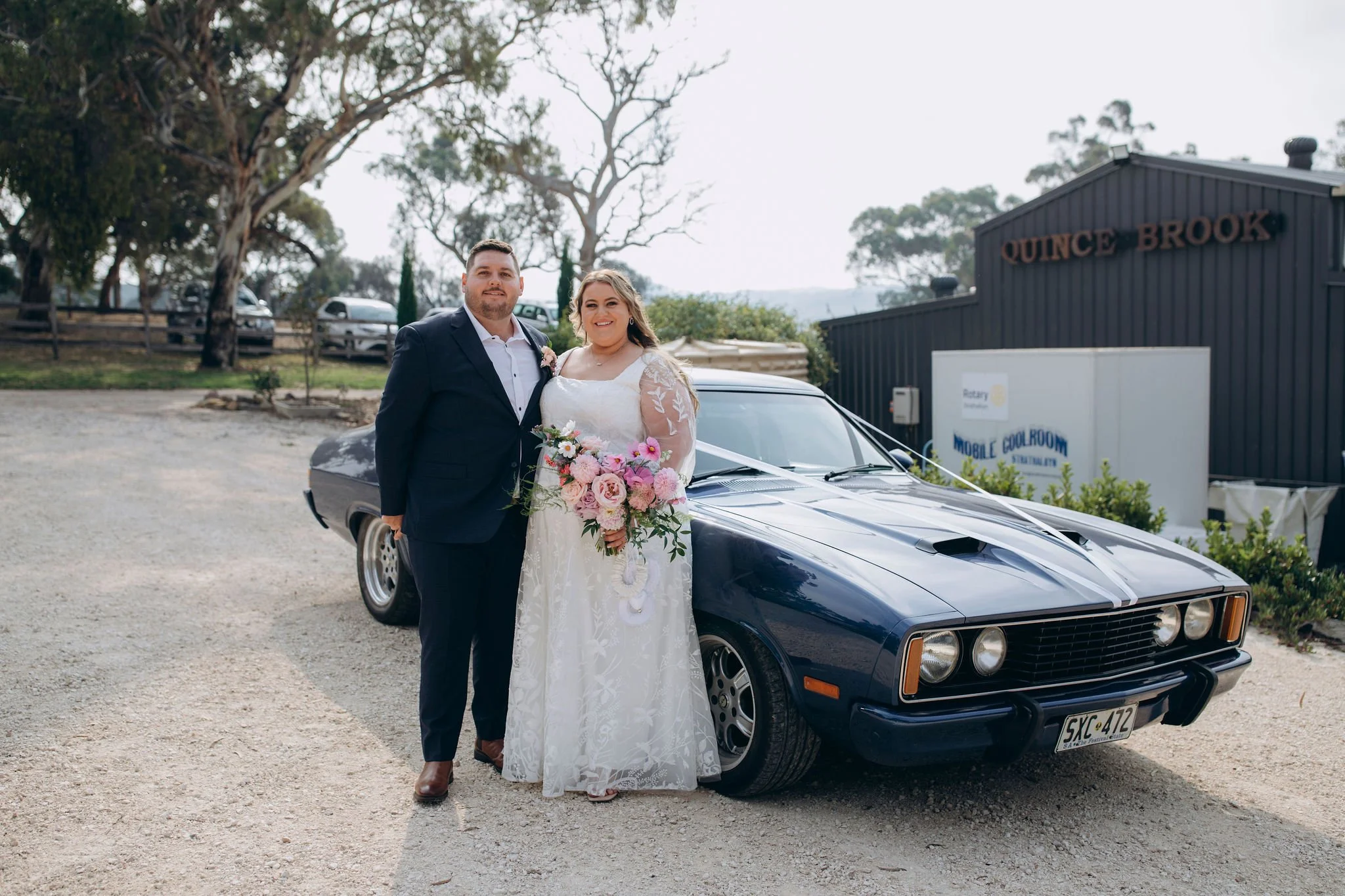 A bride and groom standing together in front of a dark blue vintage car, with the bride holding a flower bouquet, on a gravel lot with trees and a black building in the background.