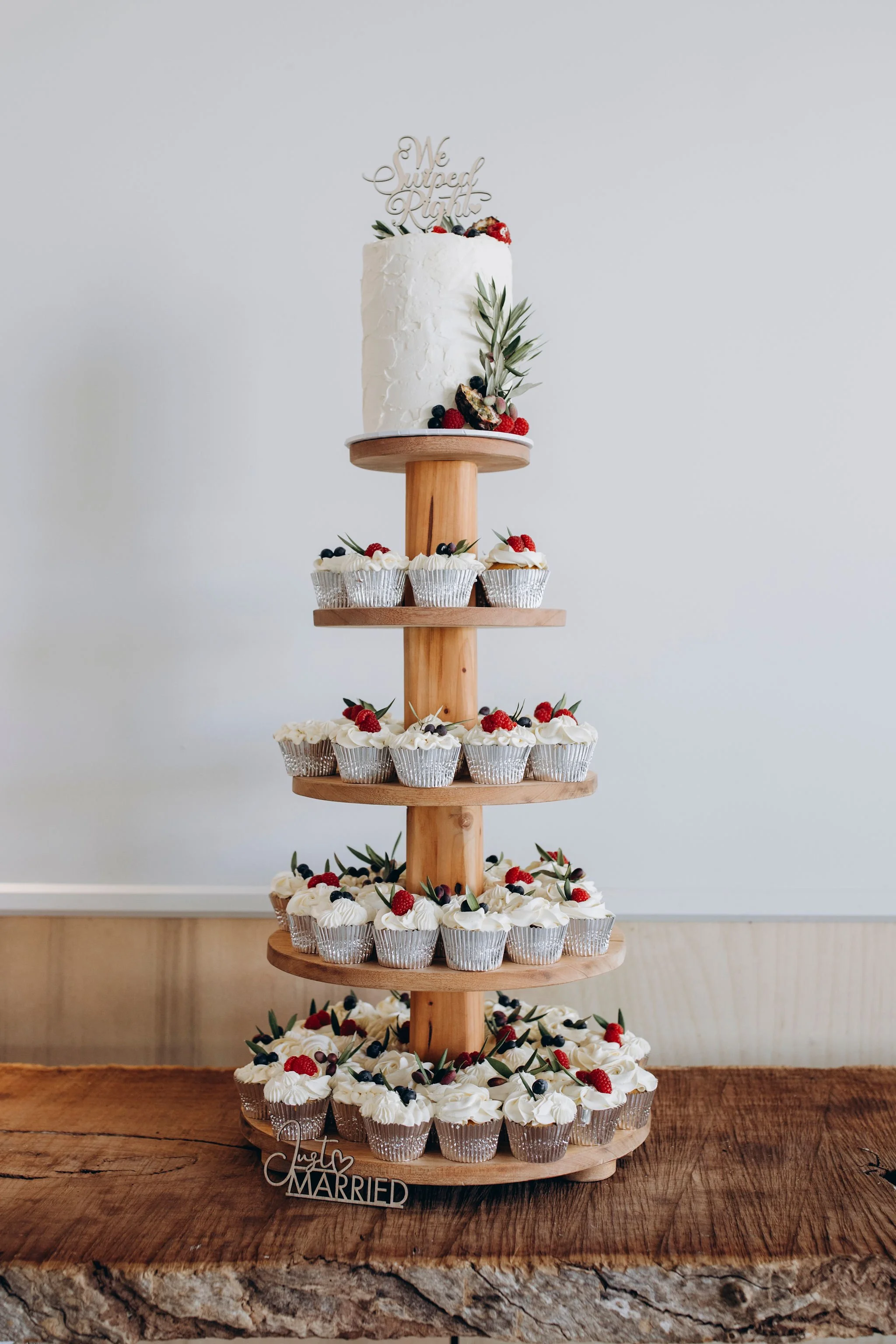 A layered dessert stand with cupcakes and a wedding cake, decorated with berries and foliage.
