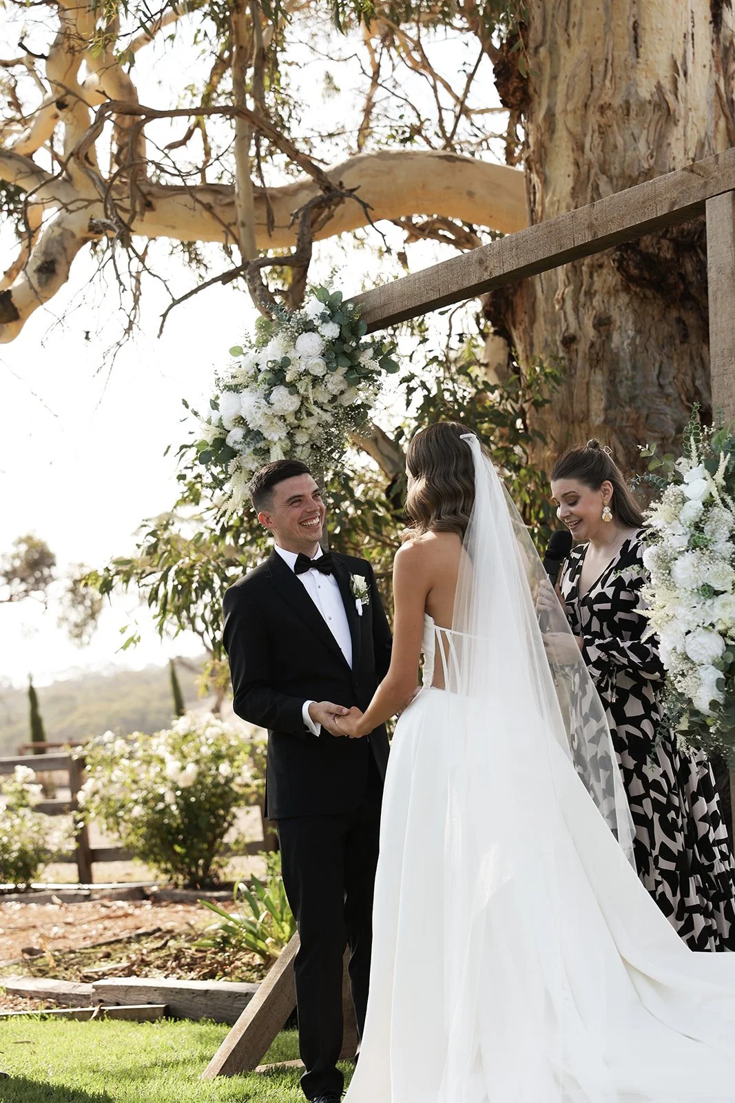 A wedding ceremony outdoors with a bride and groom holding hands, standing under a wooden arch decorated with white flowers. A woman officiant or speaker is holding a microphone, reading as the couple smiles at each other. The background features tre
