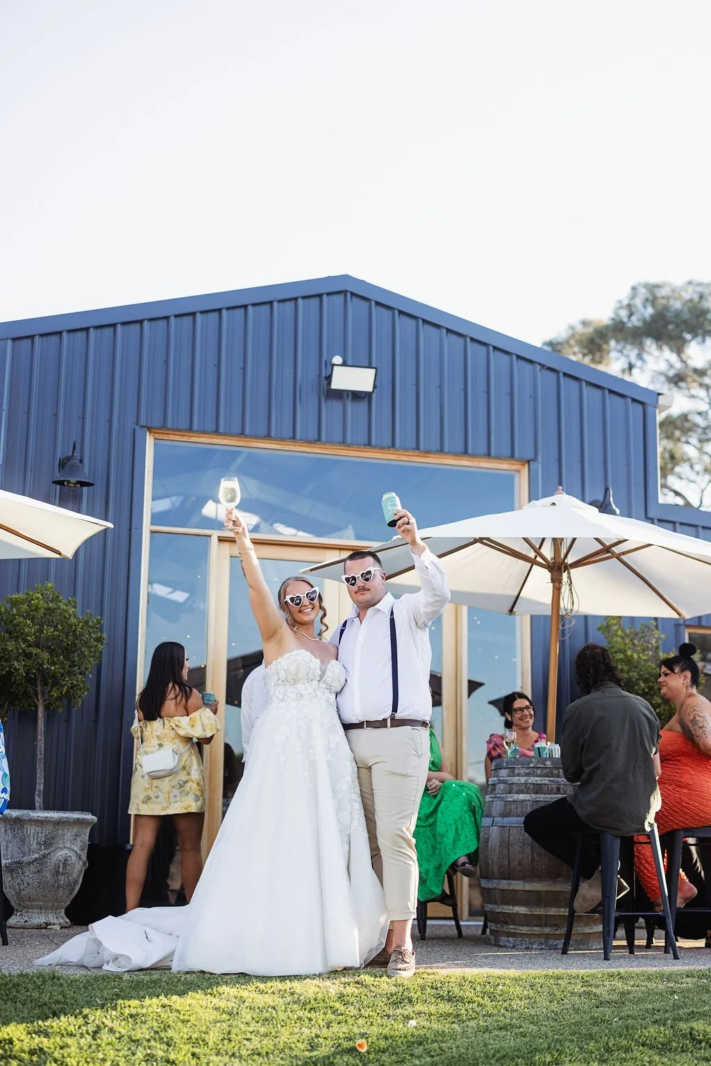 Wedding celebration with couple in wedding attire holding glasses, surrounded by friends outdoors near a blue building with large window and white umbrellas.