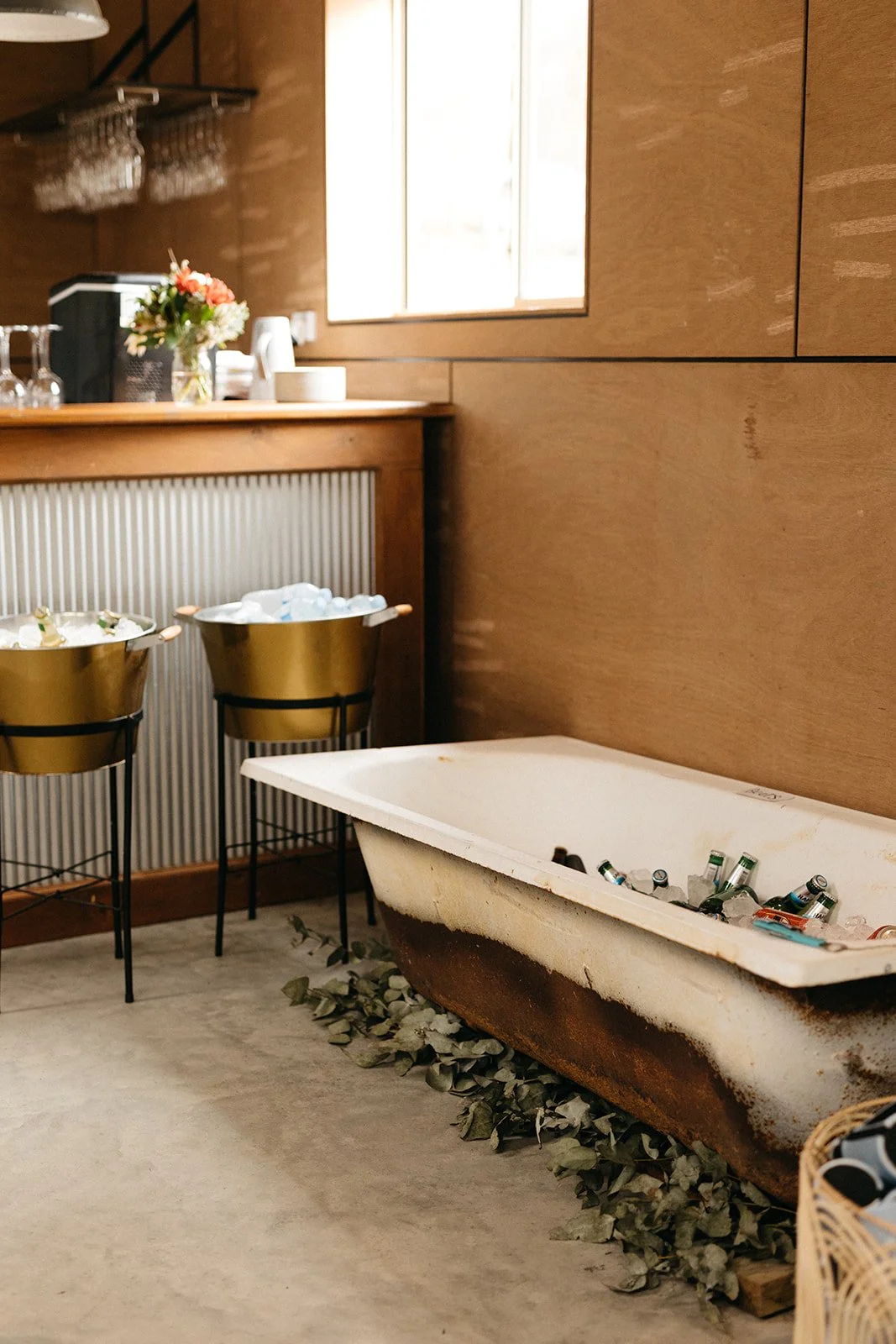 Vintage bathtub filled with bottles and cans, placed on the floor with green leaves underneath, next to a cafe bar area with gold bucket containers holding ice and bottles.