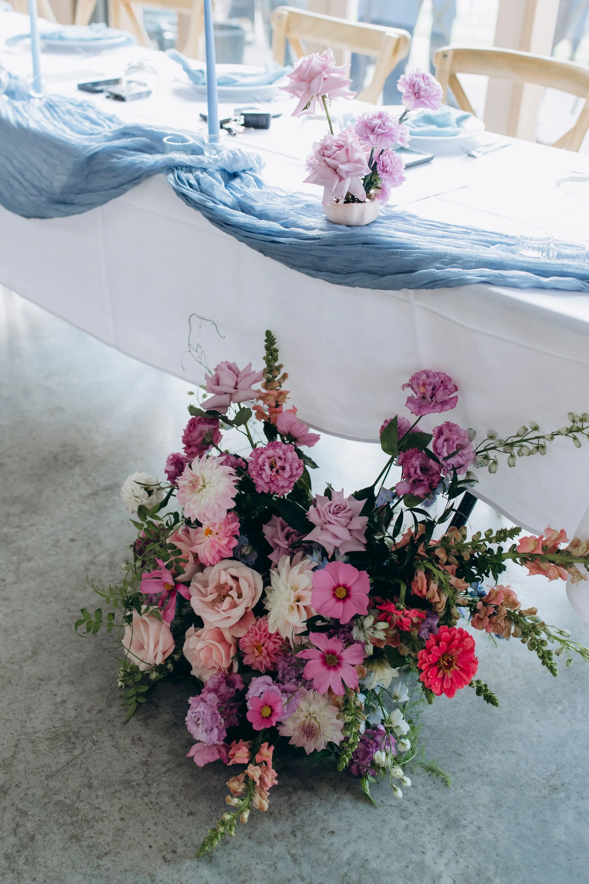 A floral arrangement with pink, purple, and white flowers on an indoor dining table with a white tablecloth and a blue fabric runner.