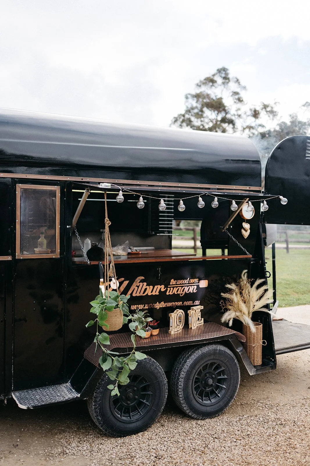 A black food truck with decorations including hanging string lights, a potted plant, and wooden letters spelling "LOVE." The truck has a sign with the name "Vilbur the wood oven wagon" and a website address. It is parked on a gravel surface with a gr