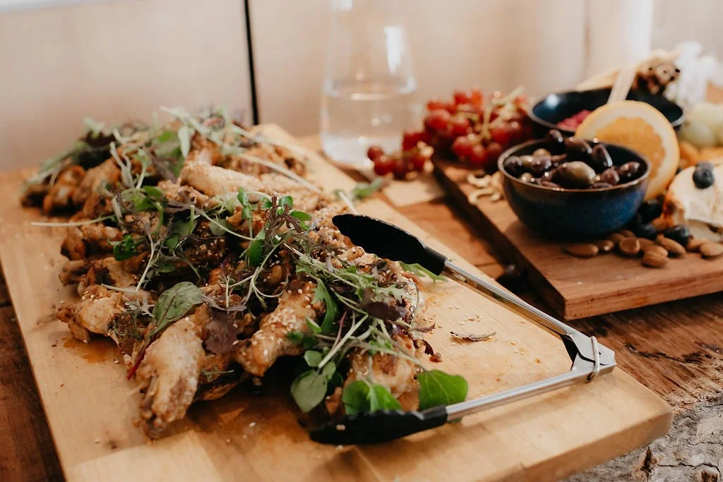 A wooden board with fried chicken wings topped with microgreens, alongside a serving utensil. In the background, there is another board with assorted fruits, olives, nuts, and cheese.
