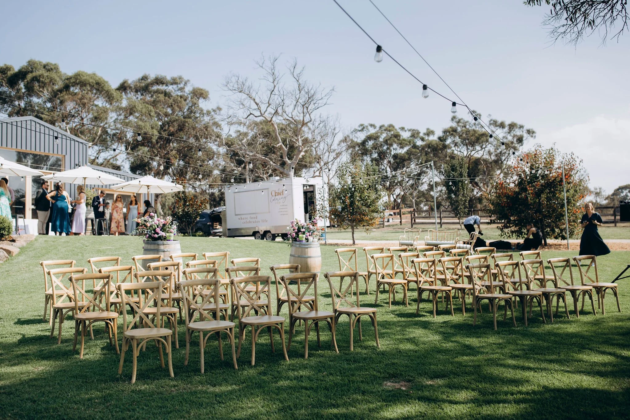 Outdoor wedding setup on a grassy area with rows of wooden chairs, flower arrangements on barrels, trees, string lights, and a food truck in the background.