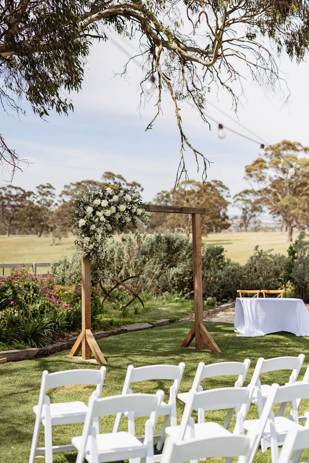 Outdoor wedding ceremony setup with white chairs arranged on a grassy area, a wooden wedding arch decorated with white flowers, a table with a white tablecloth and two chairs, and string lights hanging overhead amid trees and open fields.