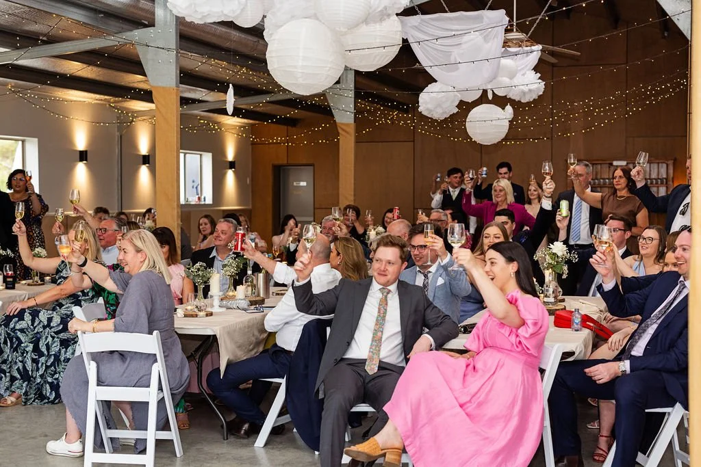 Group of guests at a celebration raising their glasses in a toast, seated at decorated tables with floral centerpieces in a warmly lit, decorated venue with hanging paper lanterns.