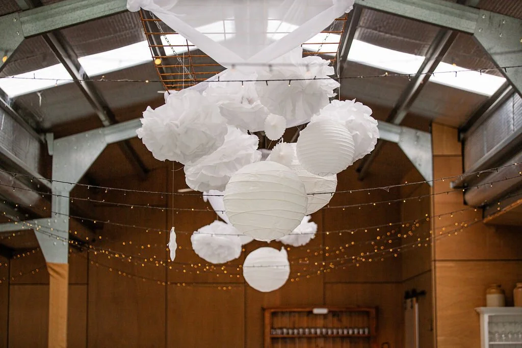 Decorative hanging paper lanterns and tissue paper clouds under a glass ceiling, with string lights in a wooden interior space.
