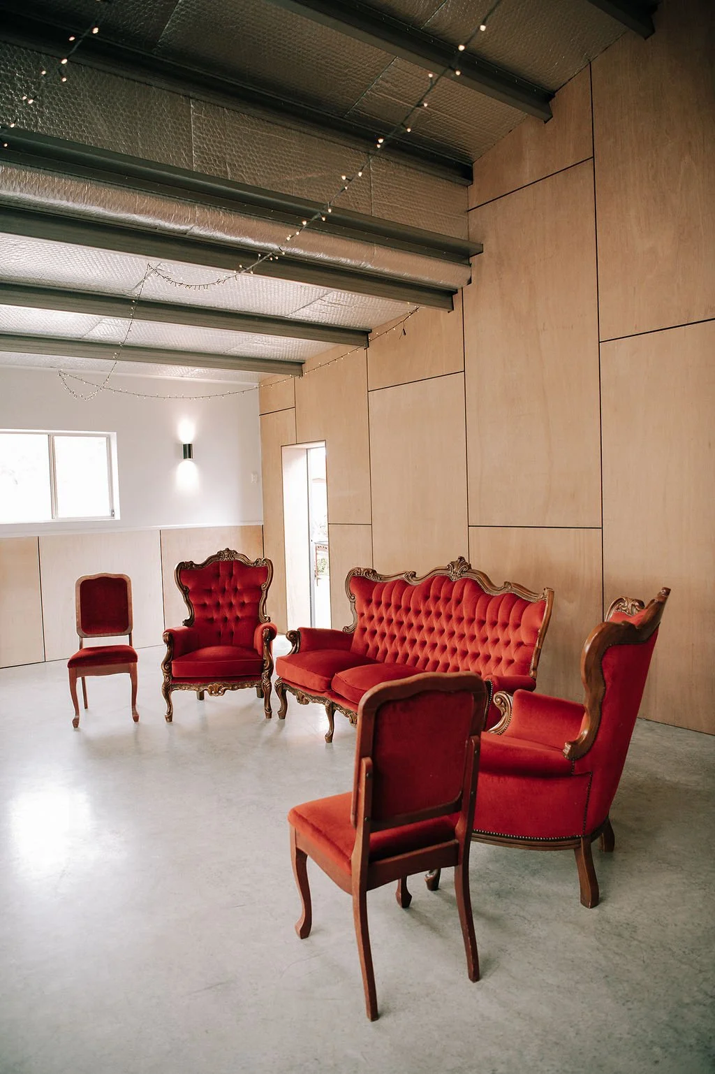 A room with six vintage red chairs, including two armchairs, a loveseat, and three side chairs, arranged on a light concrete floor against a wooden wall, with a window and string lights on the ceiling.