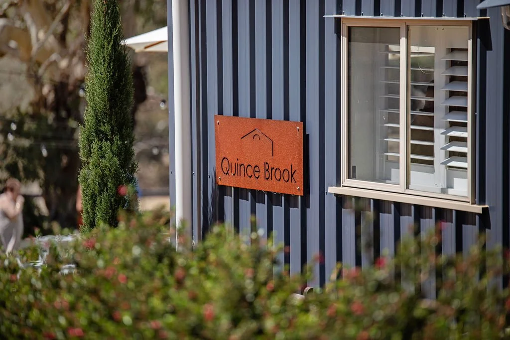 A blue corrugated metal building with a window having white shutters and a wooden frame. A rust-colored sign reads "Quince Brook" mounted on the wall. Some greenery and trees are visible in the foreground and background.