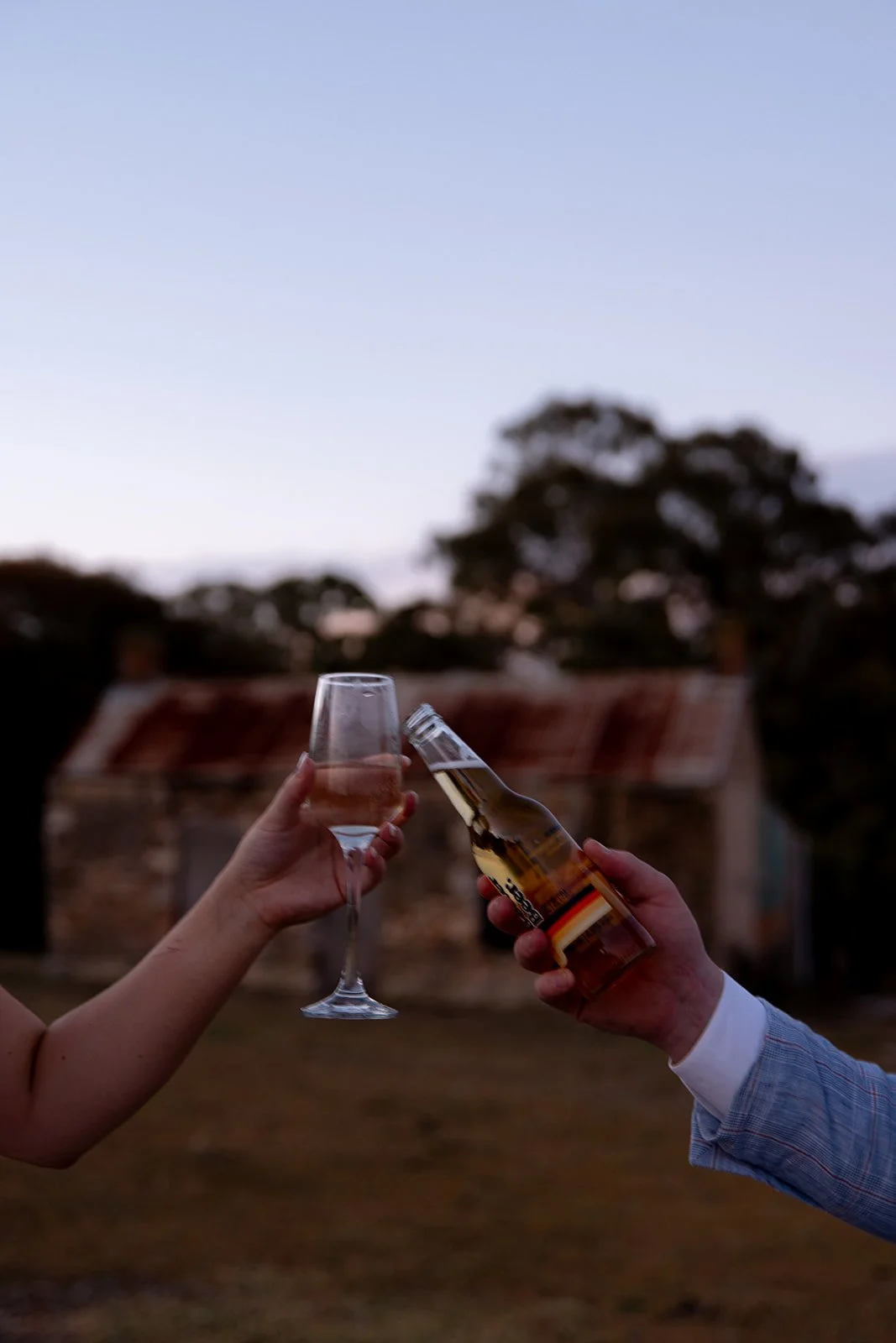 Two people clinking glasses, one with a champagne flute and the other with a beer bottle, outdoors during dusk with trees and an old building in the background.
