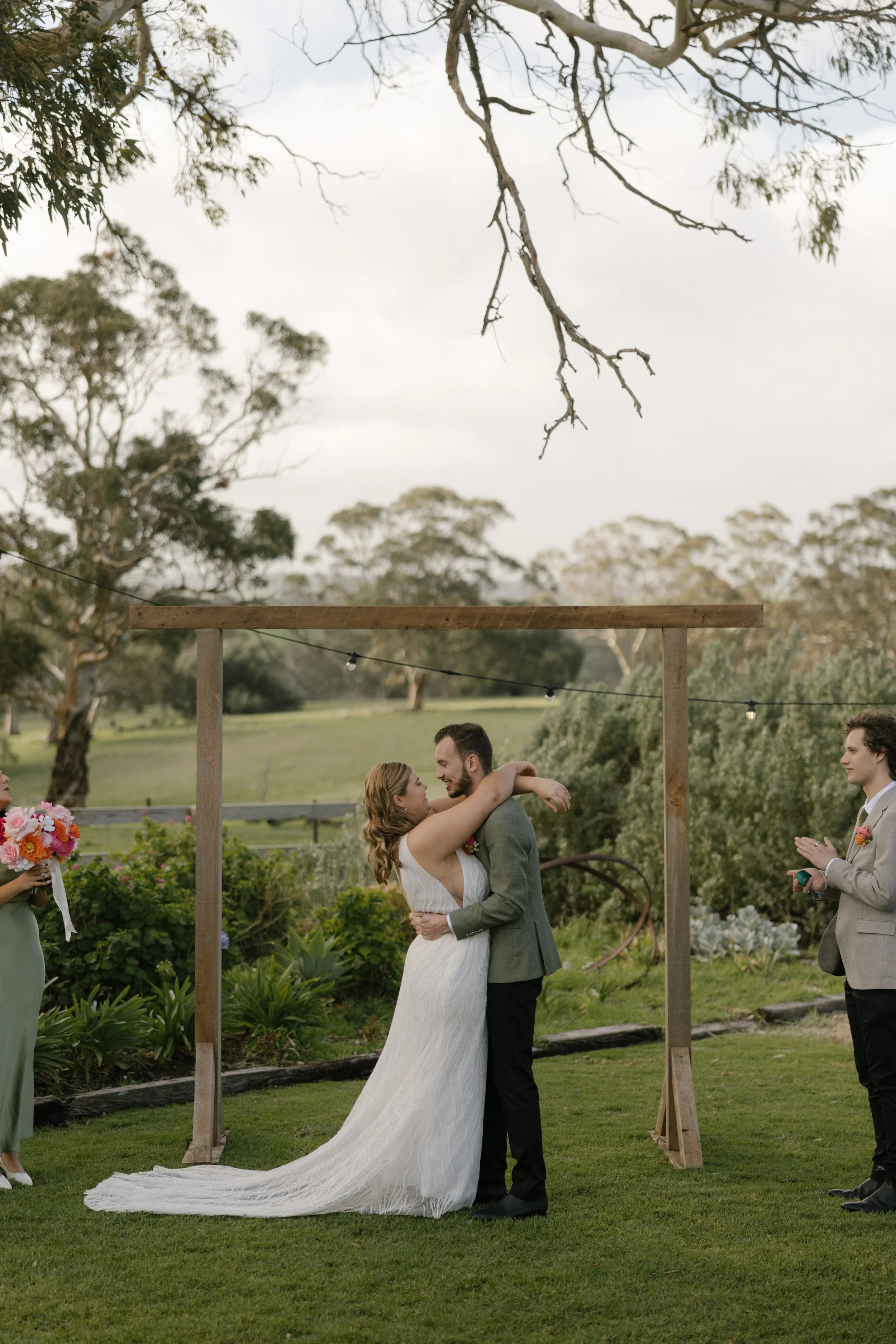A couple in wedding attire hugging during an outdoor wedding ceremony, surrounded by greenery and trees, with an officiant and bridesmaid holding bouquet in the background.