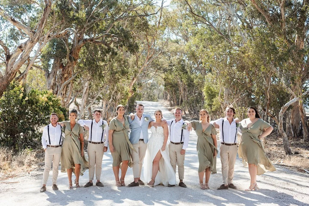 A bride and groom with bridesmaids and groomsmen standing on a dirt road surrounded by trees.