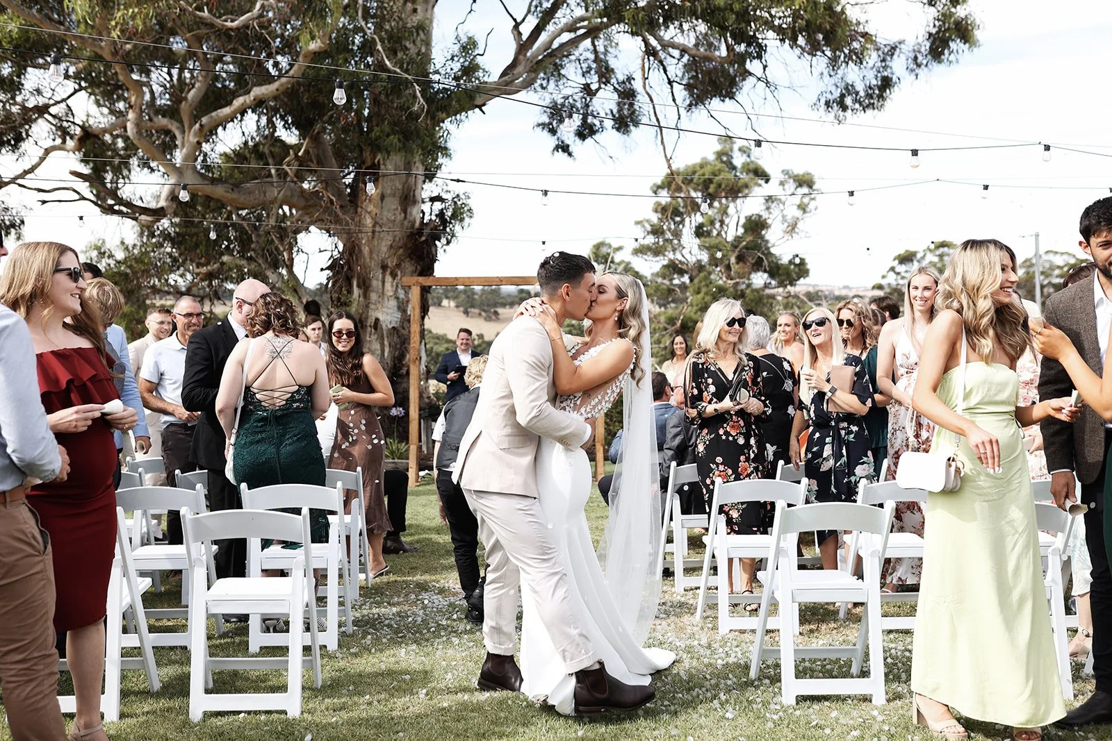 A wedding ceremony outdoors with a newlywed couple kissing amid guests, under string lights, large trees, and a scenic landscape in the background.
